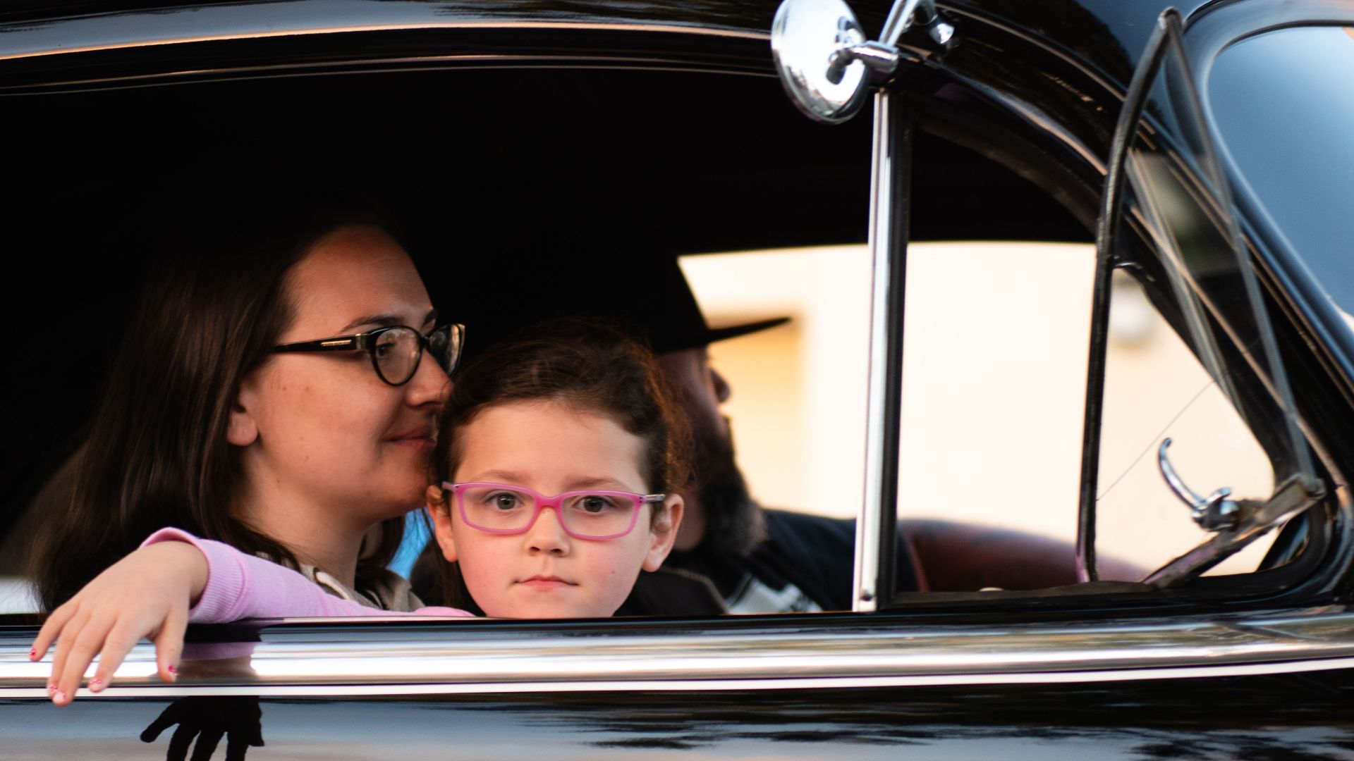 A mother and daughter enjoy a car ride in a vintage black automobile, capturing a serene family moment.