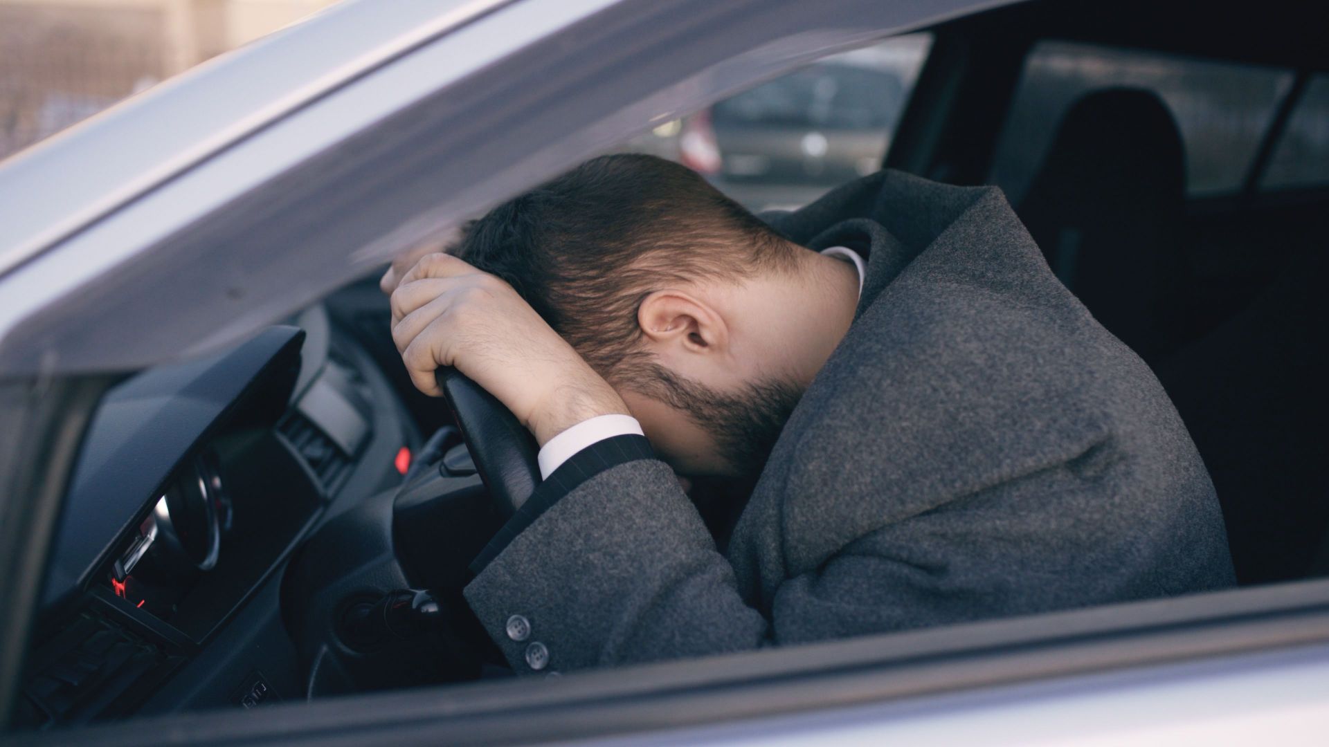 Man feeling overwhelmed and stressed, resting his head on a steering wheel inside a car.