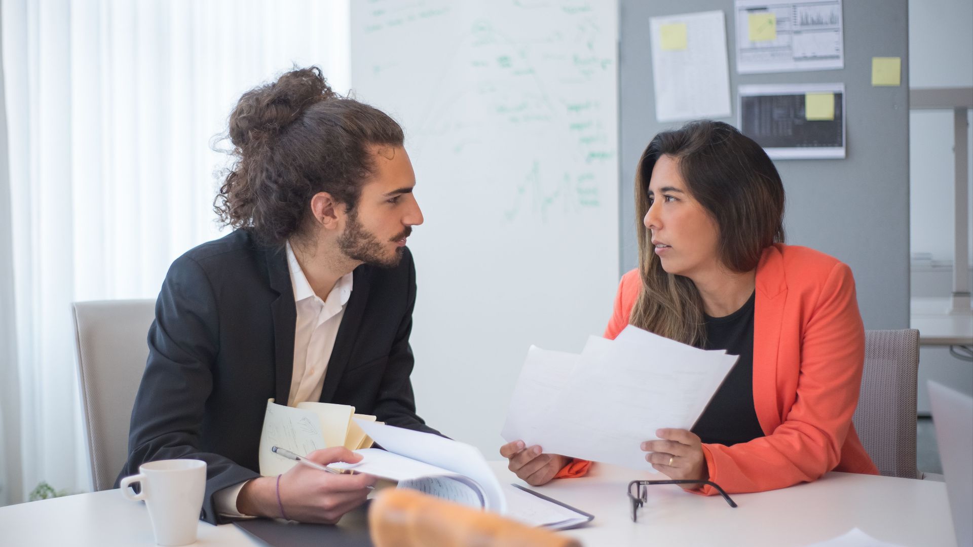 Two colleagues engaged in a discussion at an office table with documents and coffee.
