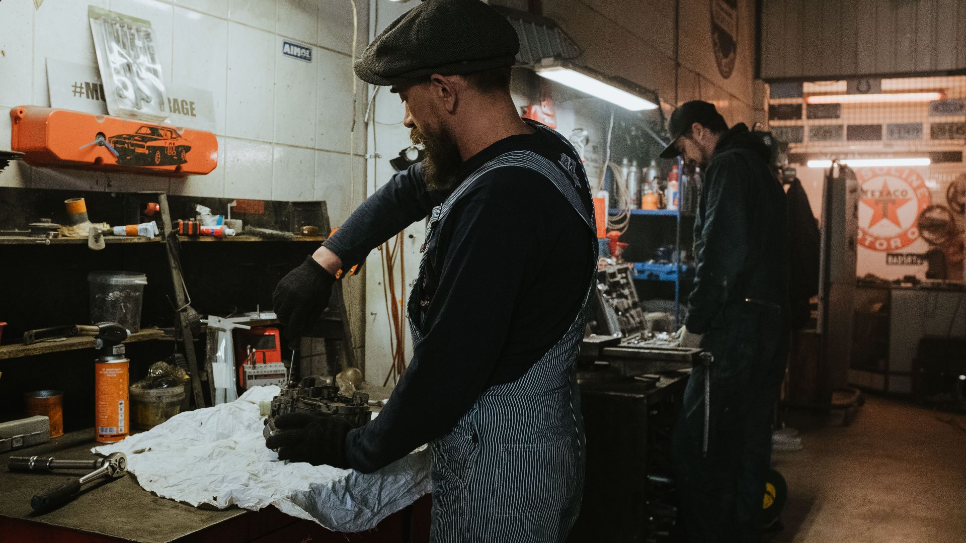 Two auto mechanics engaged in vehicle repair work inside a dimly lit garage.