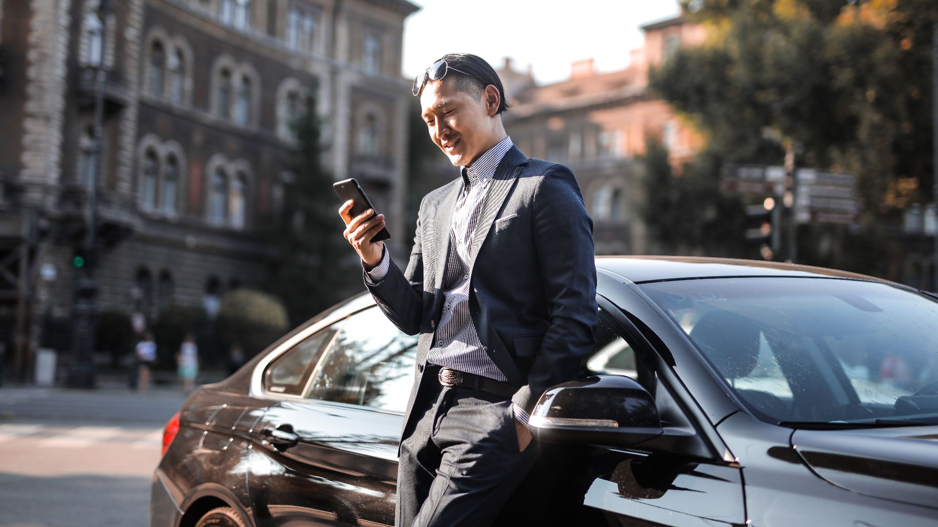 Smiling businessman in a black suit texting on his phone while leaning against a parked car in the city.