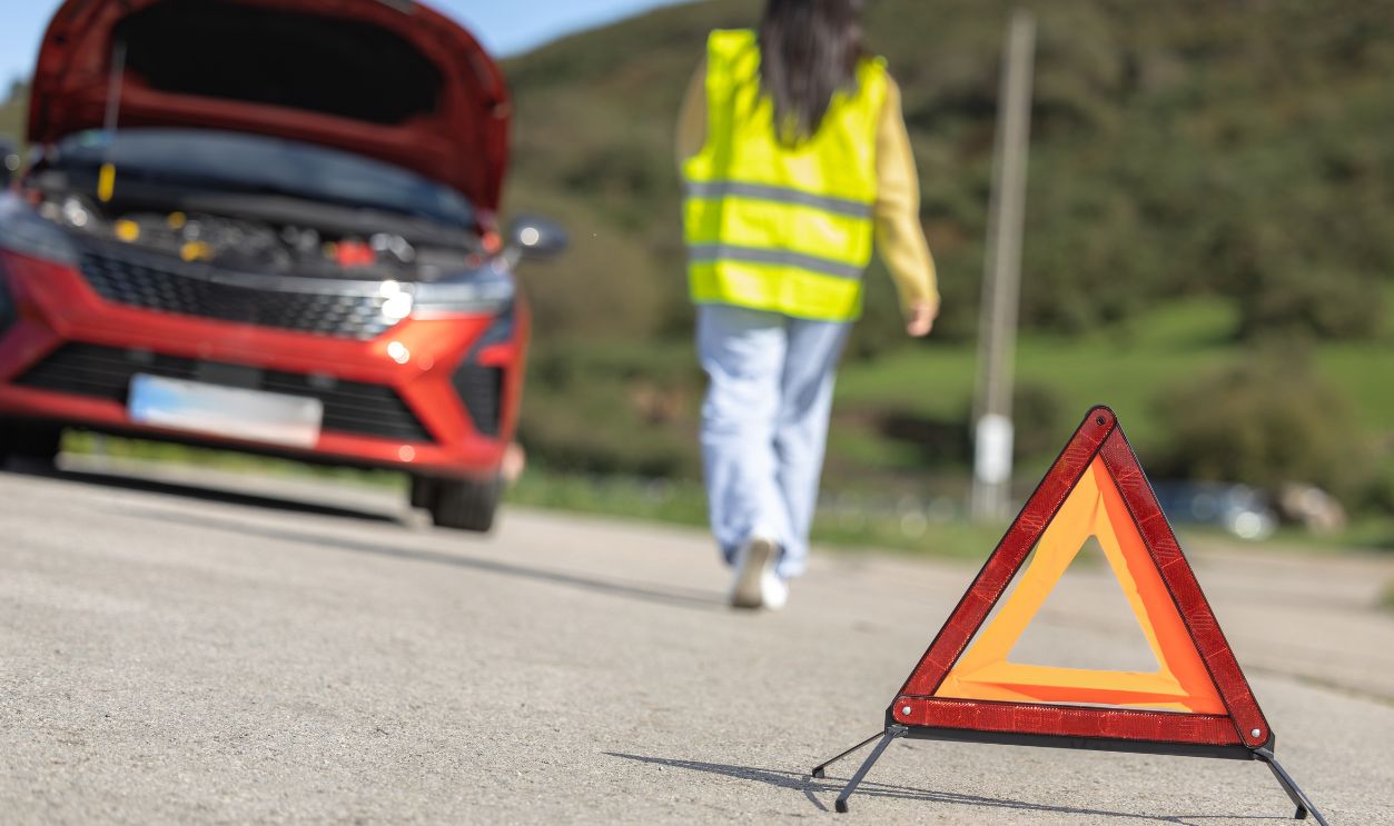 Pretty young woman poses in closeup placing emergency triangle sign and reflective vest on the road, broken car, roadside assistance, waiting for insurance tow truck