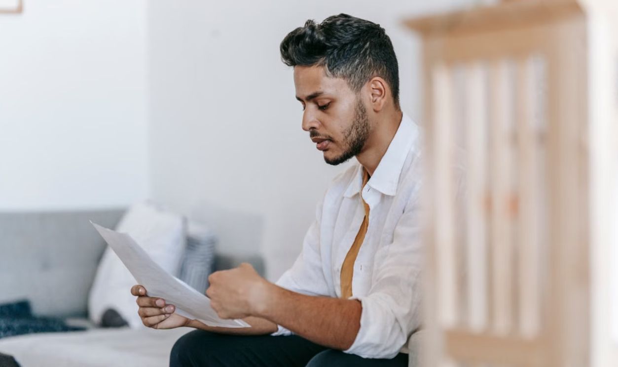 Young man in a white shirt reading papers at home, appearing thoughtful and focused