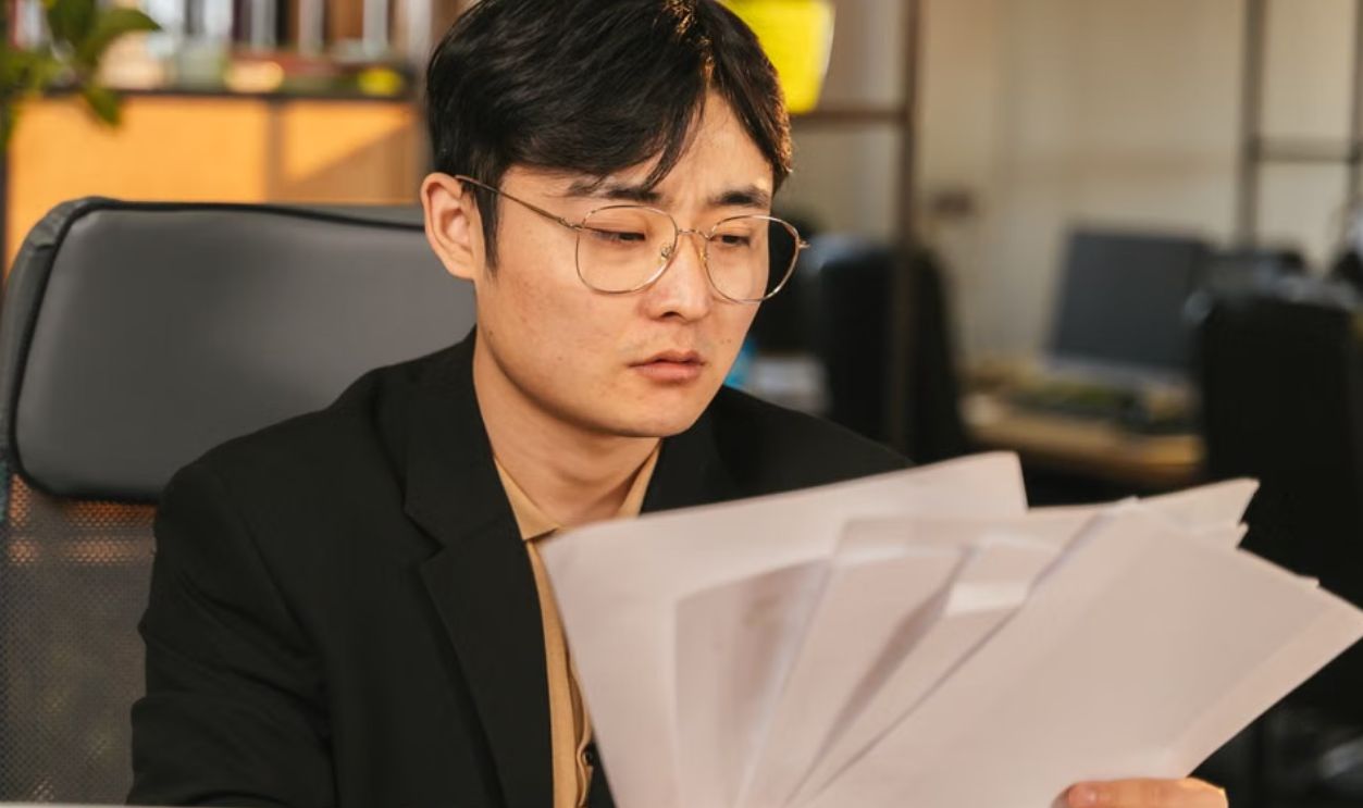 Asian man in glasses and suit analyzing papers at his desk in a modern office