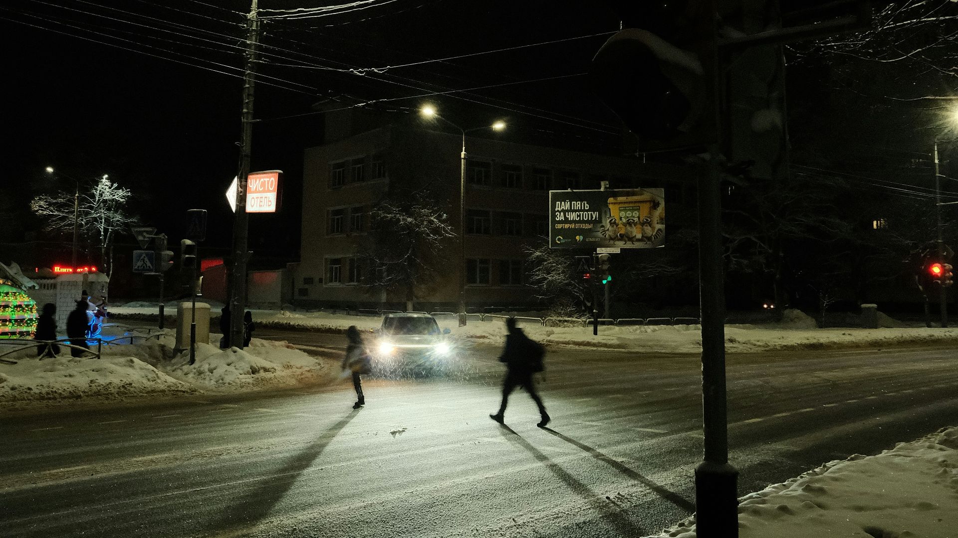 Pedestrians cross a snowy street at night with traffic light.