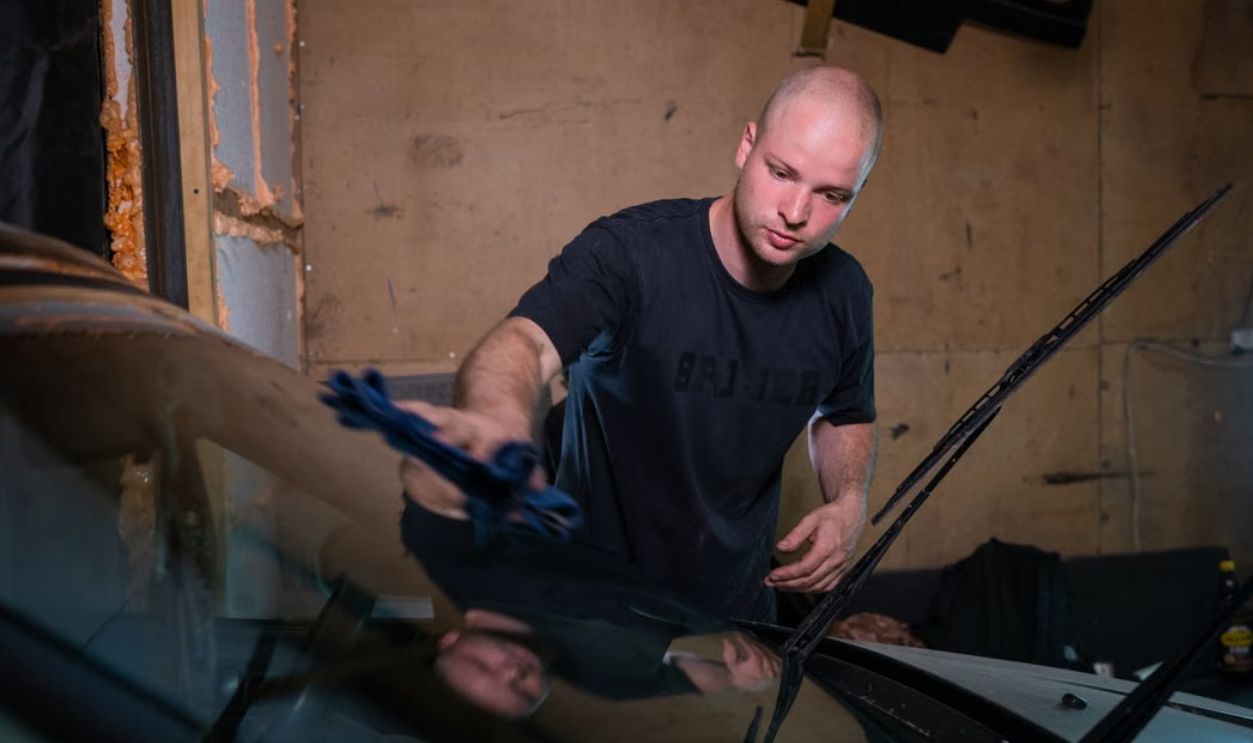 A man wipes a car windshield inside a garage, focused on cleaning