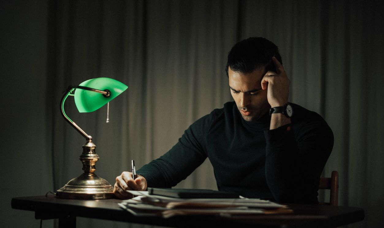 Focused man working late at a desk, lit by a characteristic green banker’s lamp, surrounded by paperwork