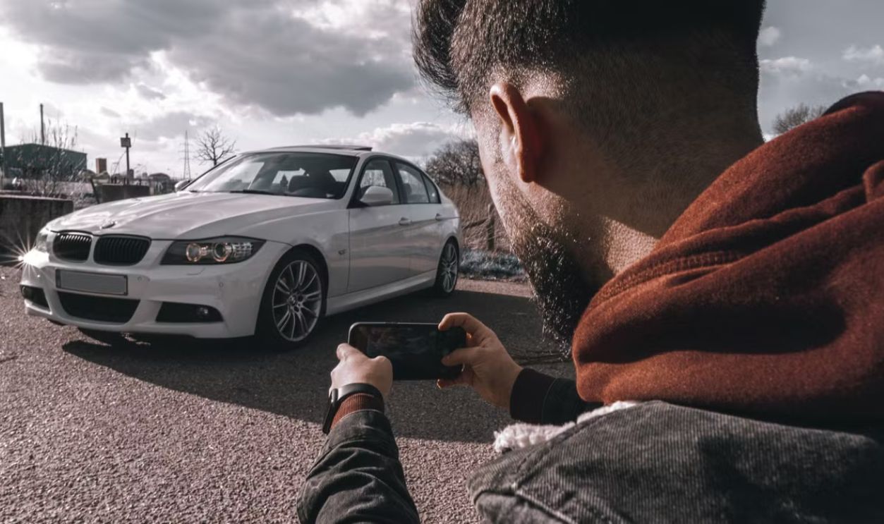 A man takes a picture of a white luxury car using a smartphone outdoors under a cloudy sky