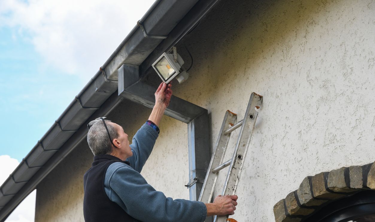 Electrician reaches for motion sensor LED floodlight while standing on ladder installing it on facade of house under gutter to improve outdoor lighting
