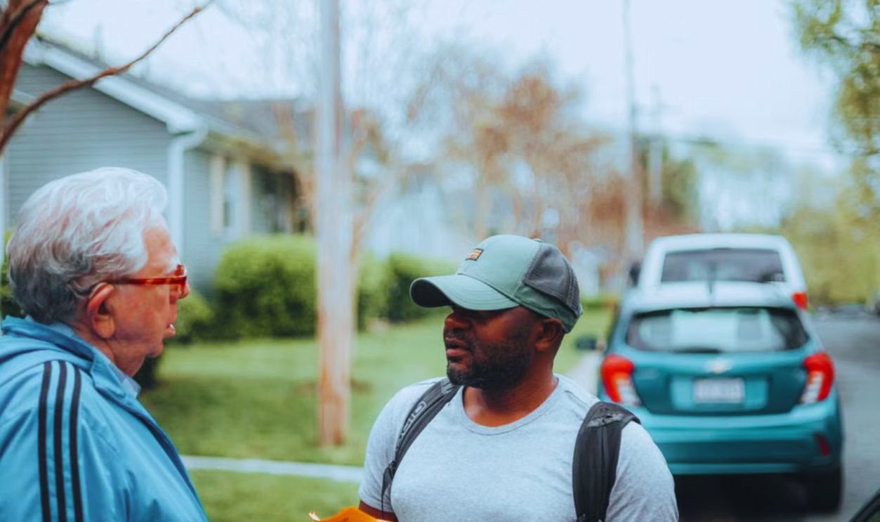 Two men engaged in conversation on a suburban sidewalk with parked cars