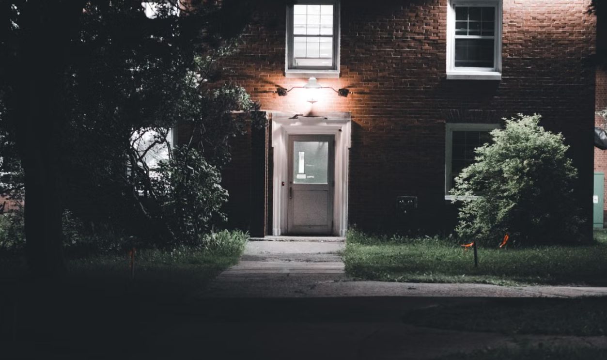A warmly lit brick house entrance under a night sky, featuring a serene garden setting