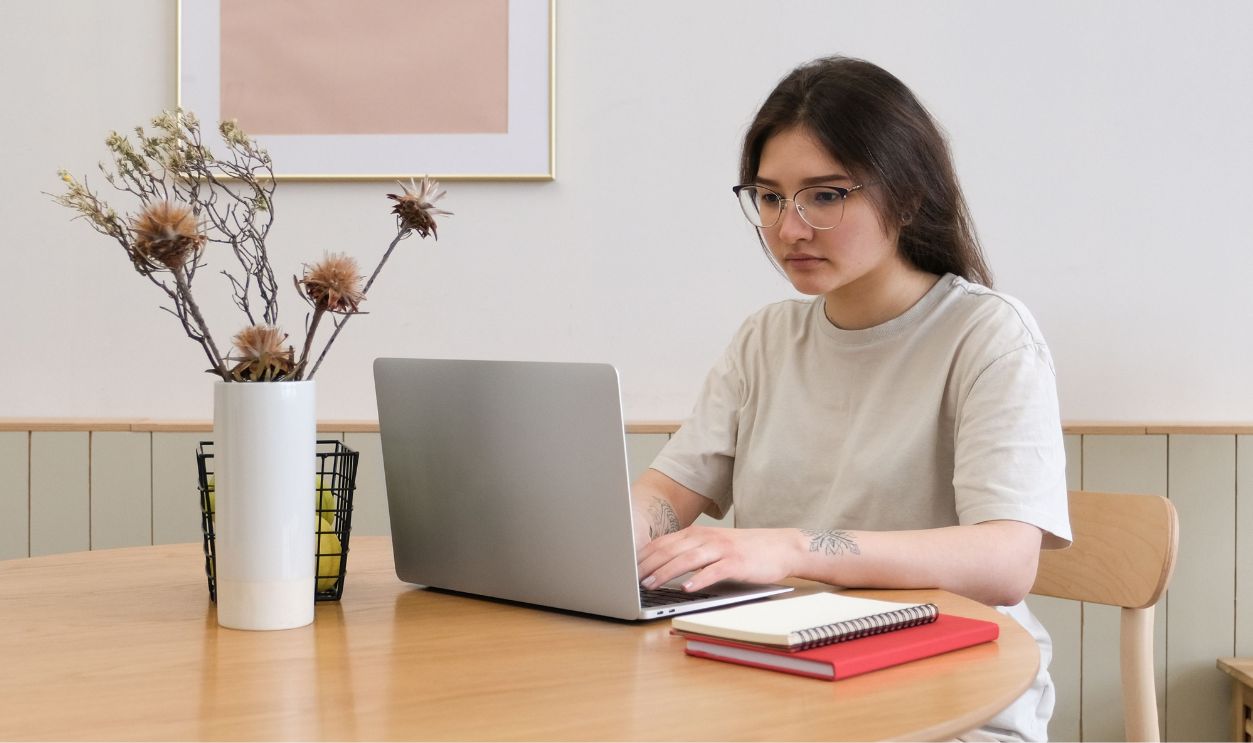 Woman sitting at a table working on a laptop from home, demonstrating a remote work setup with a vase