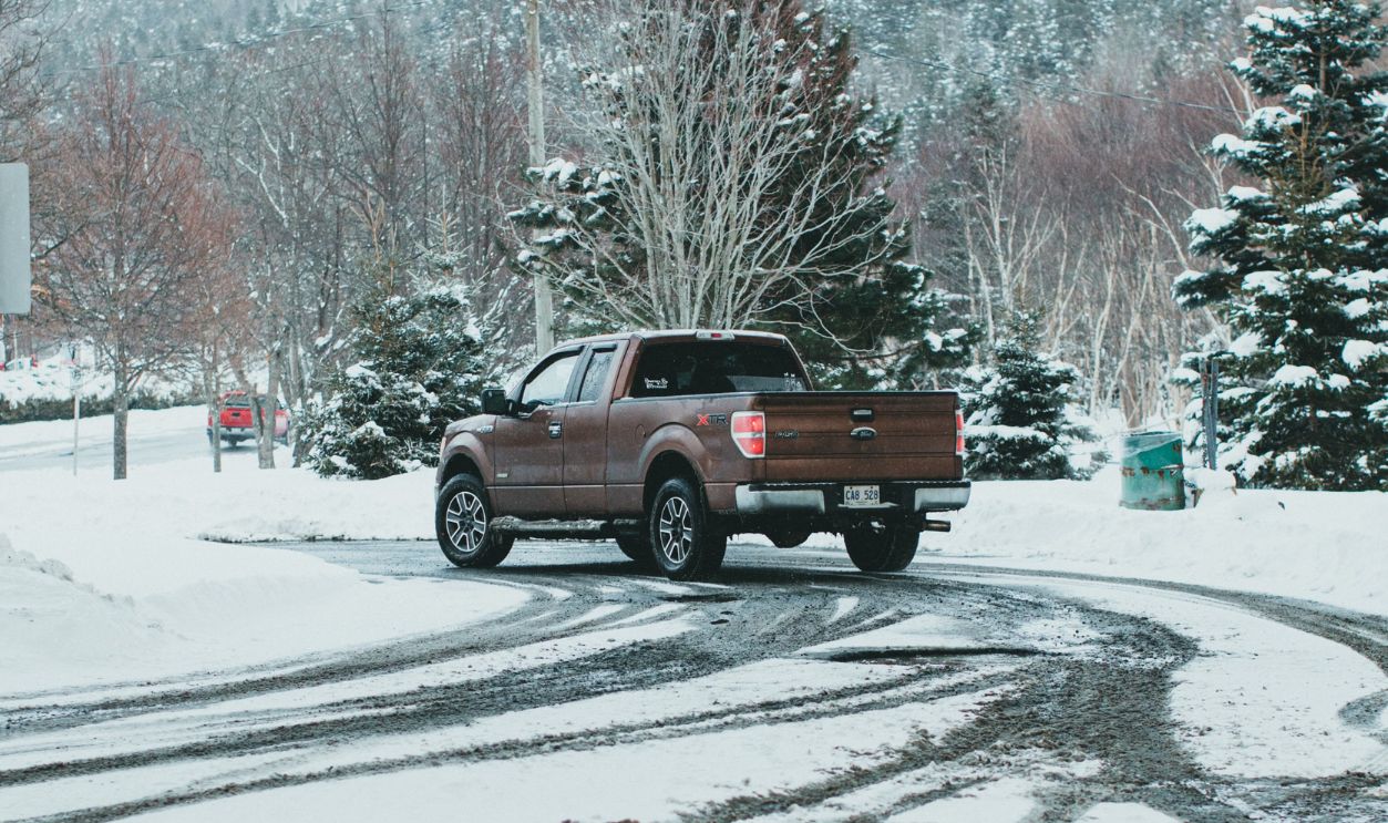 A pick-up truck navigates snowy roads in a serene winter forest, surrounded by snow-covered trees and distant hills.