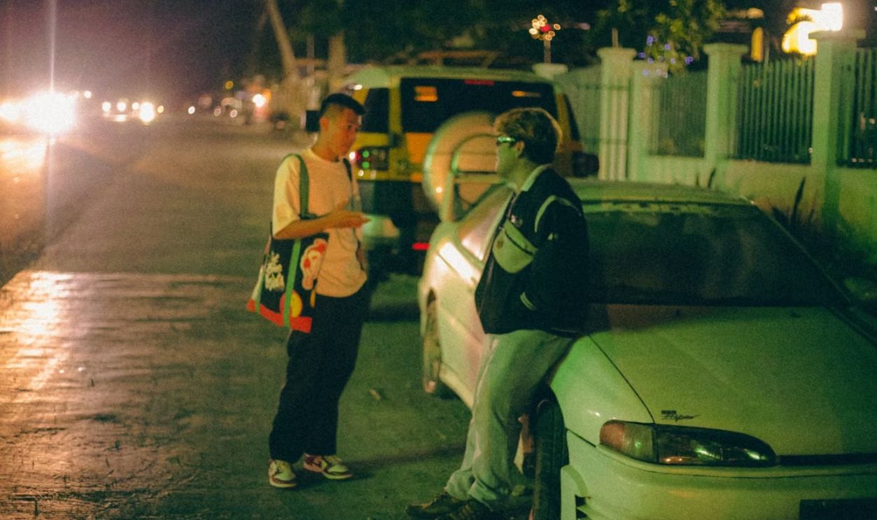 Two young men talking near a car on a dimly lit urban street at night.