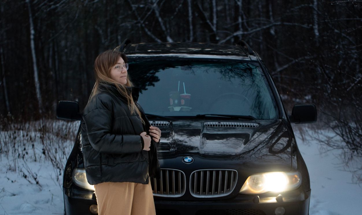 A woman stands by her BMW in a snow-covered forest, showcasing winter adventure