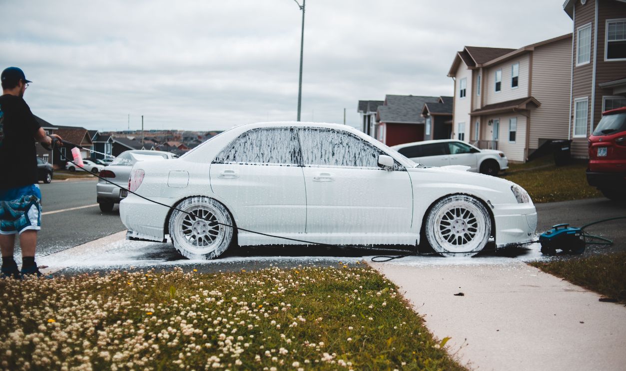 A man pressure-washes a car with foam in a suburban neighborhood