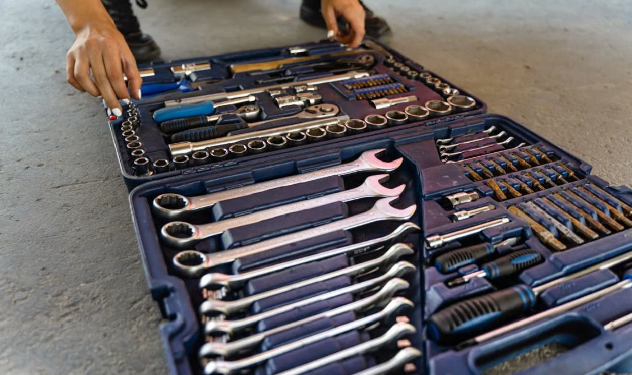 Close-up view of an open toolbox on a concrete floor, displaying various hand tools including wrenches, screwdrivers, and socket sets.