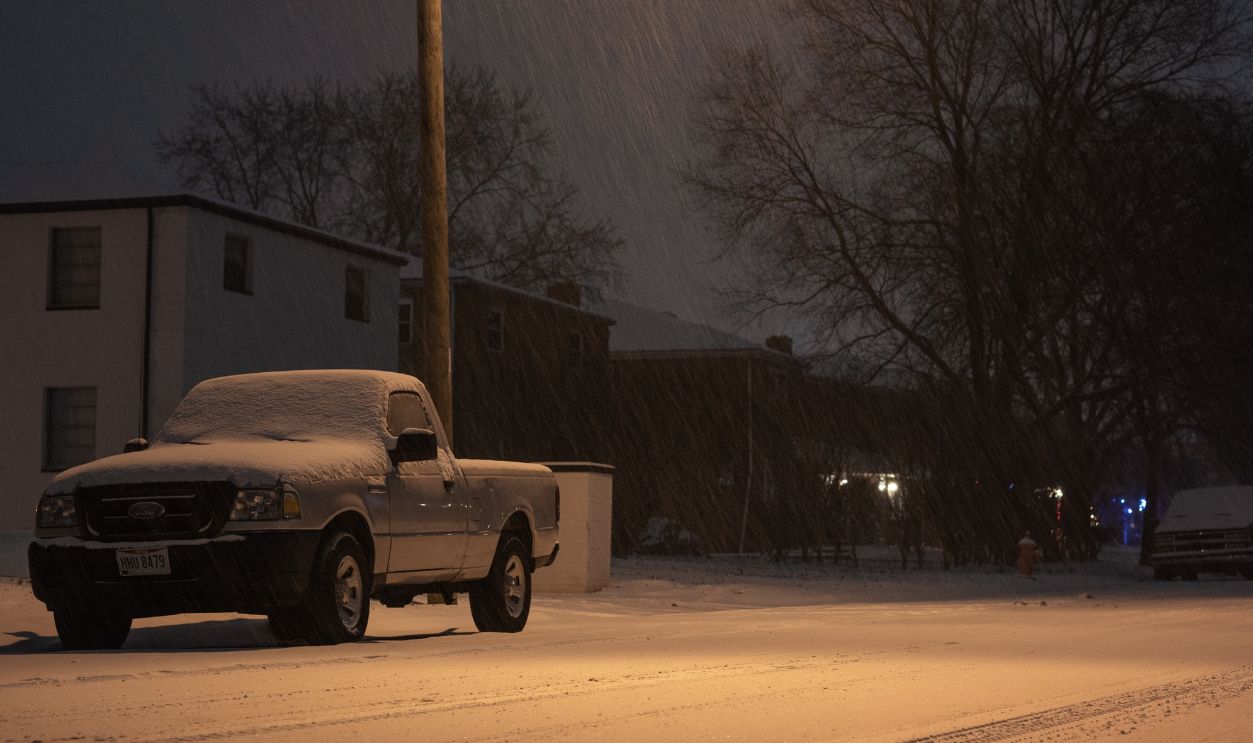 A parked pickup truck under a glowing streetlight amidst snowfall on a snowy urban street at night