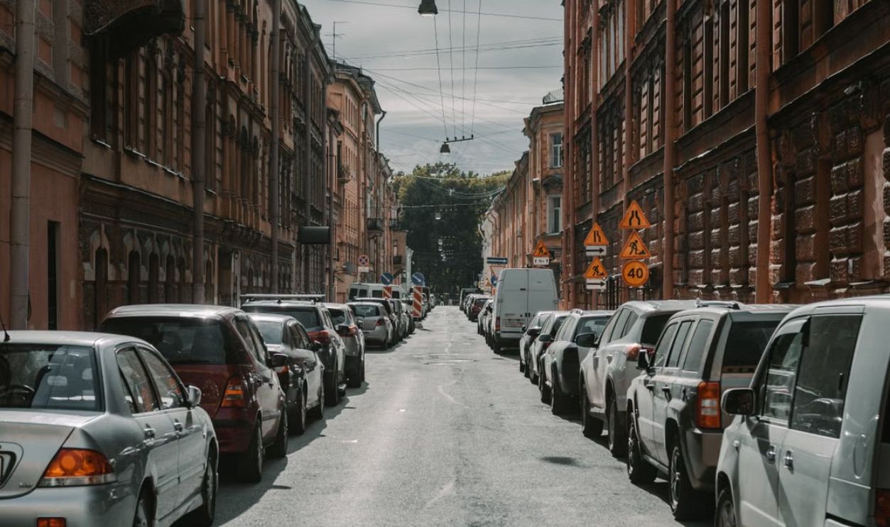 A narrow historic street lined with parked cars and classic buildings under a cloudy sky.