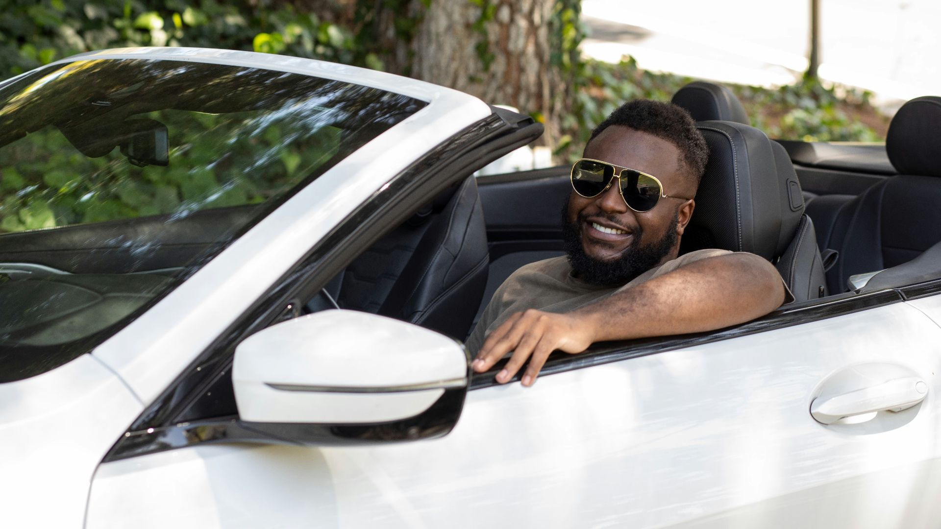 A smiling man in sunglasses drives a white convertible car.