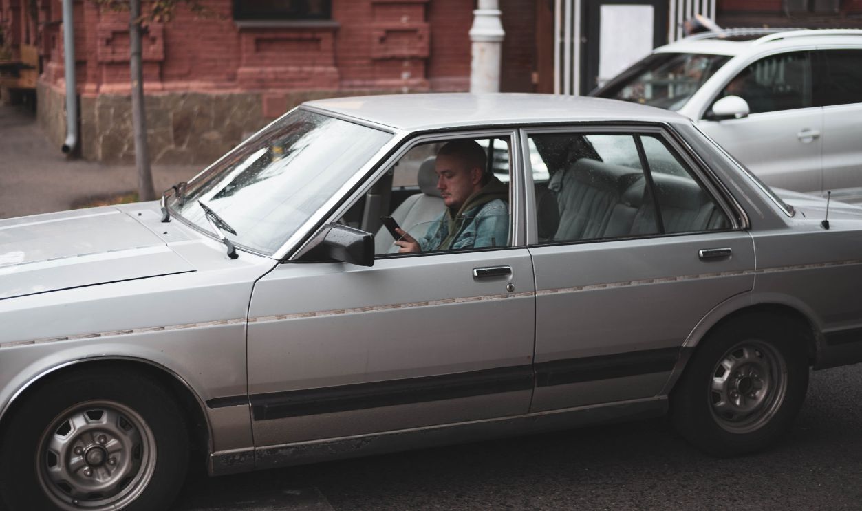 A classic silver sedan with a driver using a mobile phone in urban traffic