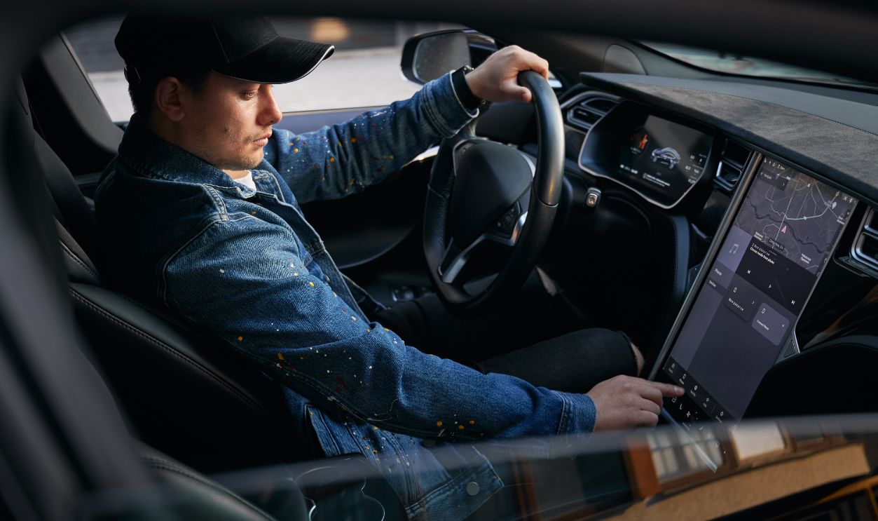 View through open side electric car window. Man in cap sitting in elite automobile, putting one hand on steering wheel and by other hand entering destination address on large touch screen.