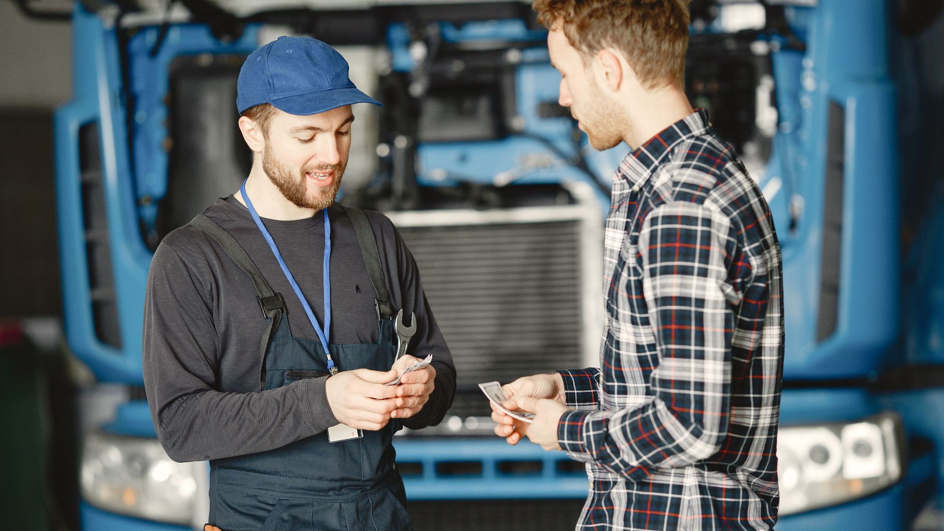 Auto mechanic in blue uniform receives payment from customer in a garage setting with a truck in the background.
