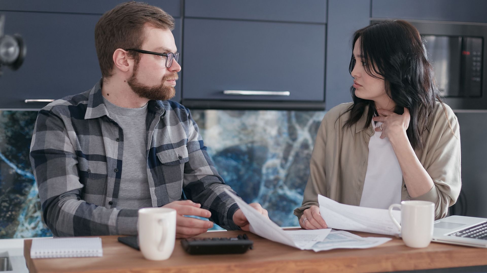 A young couple sitting at a table discussing bills and financial plans in a modern kitchen.