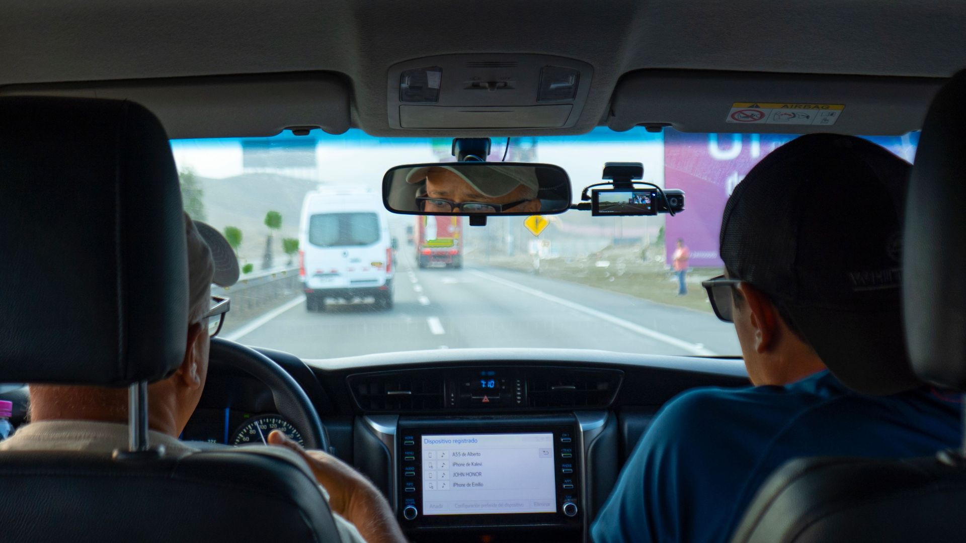 Interior of a car with two people driving on a highway, focusing on dashboard and rear view mirror.