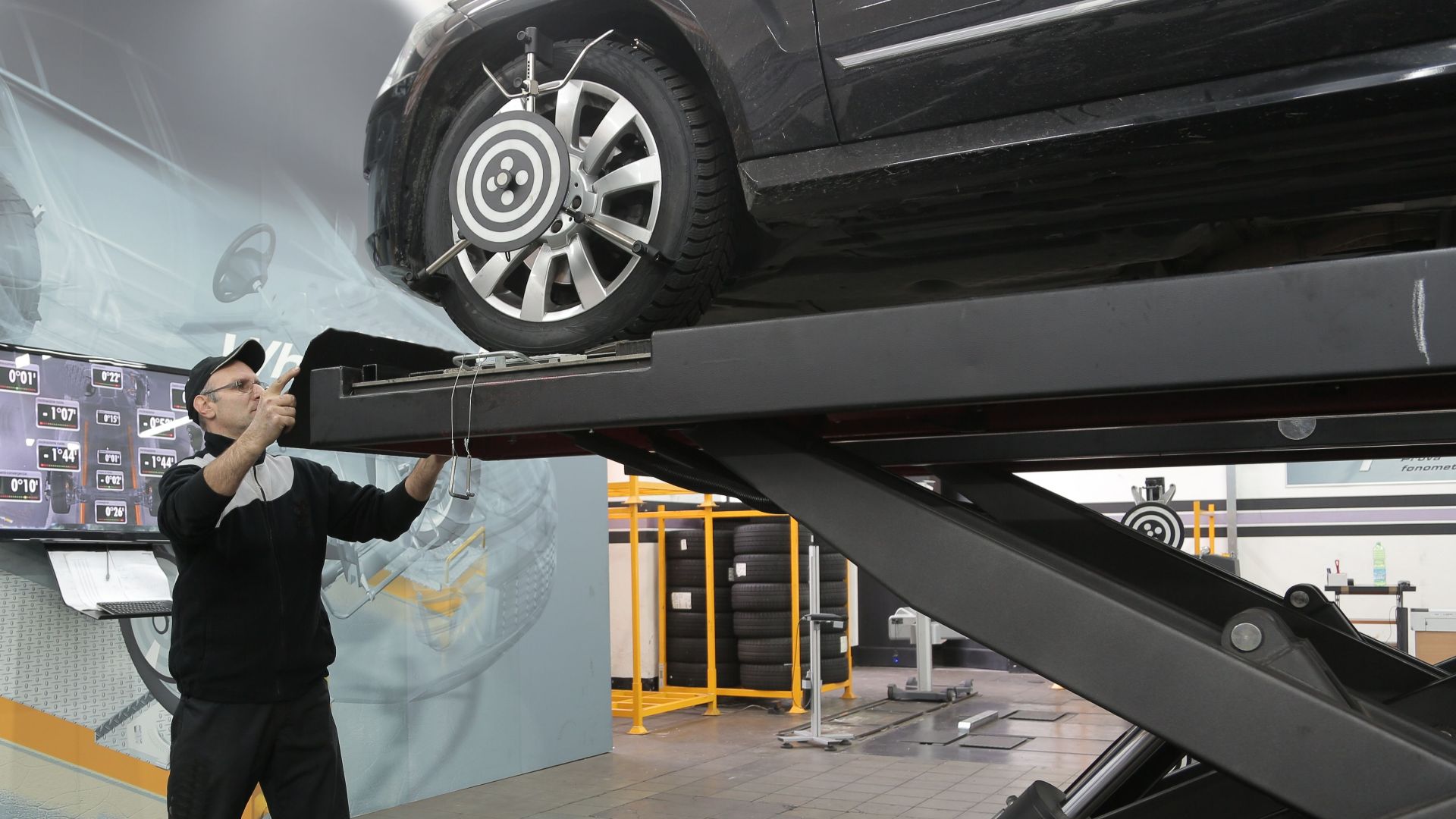 Serious professional adult mechanic in casual uniform examining car wheels while standing near lift in modern car service garage