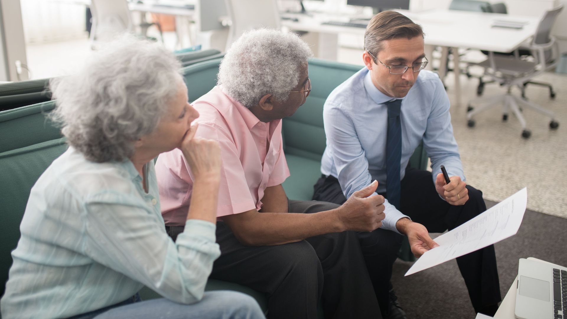 Business professional consults elderly clients in an office setting. Collaborative discussion, paperwork visible.