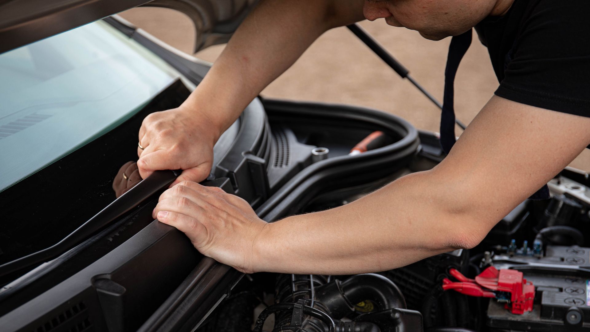 Auto mechanic working on a car engine bay, hands holding parts, outdoor setting.