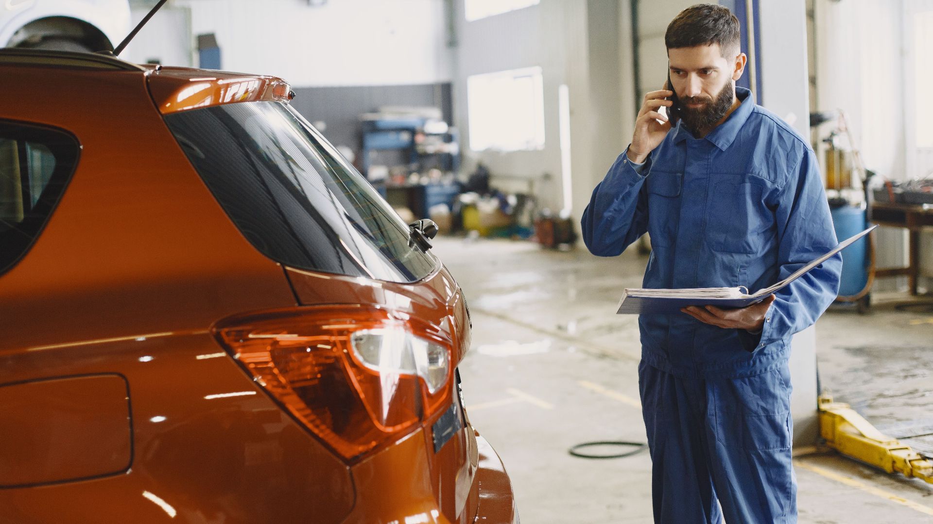 Mechanic in a blue uniform talks on phone beside a red car in an auto repair shop.