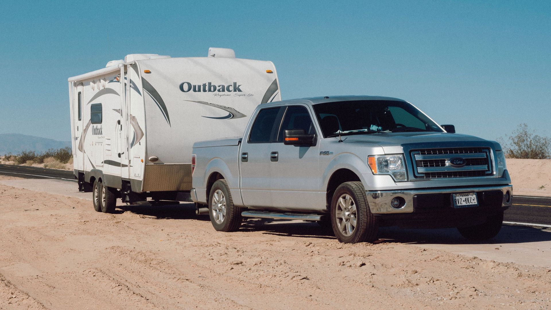 Silver truck towing an Outback trailer on a desert road under clear blue skies.