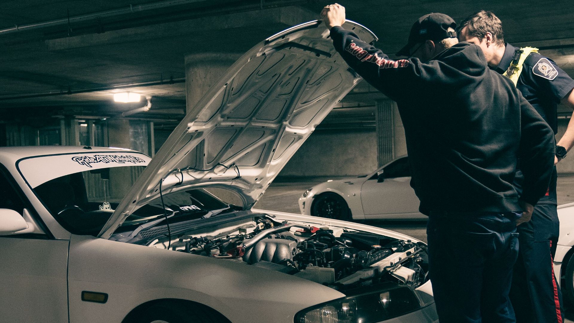 Two men examining a car engine in a dimly lit underground parking area.