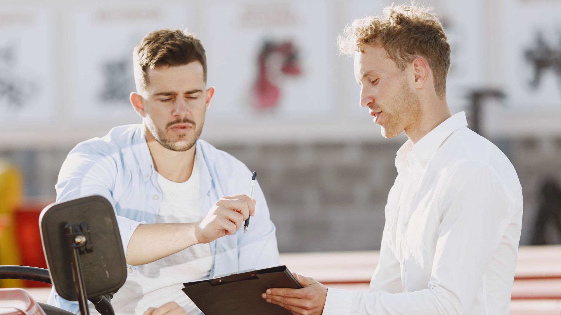 Two professionals discuss on a red tractor, examining paperwork under bright sunlight.