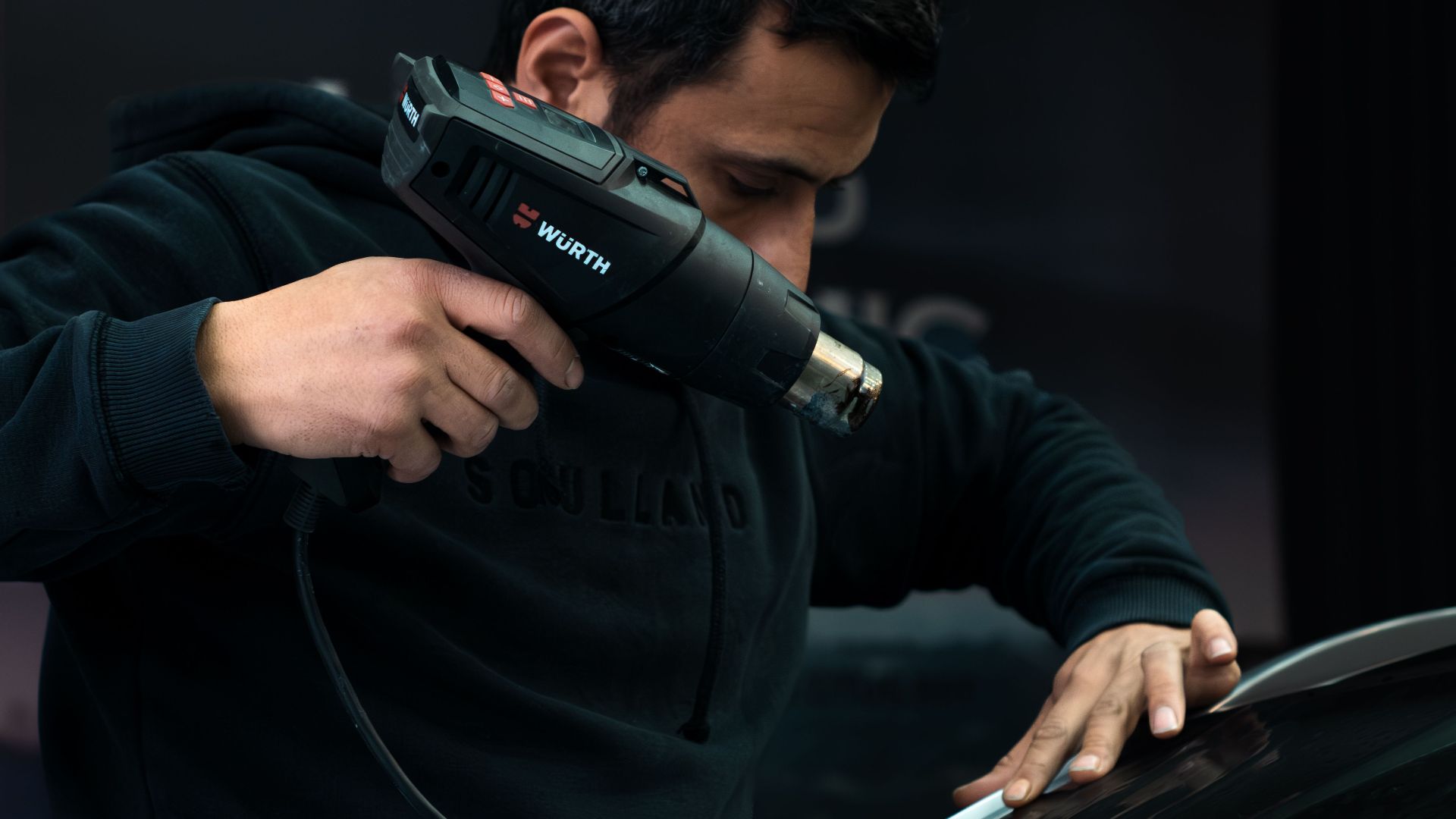 Mechanic carefully applies heat to car windshield with a heat gun in a garage setting.