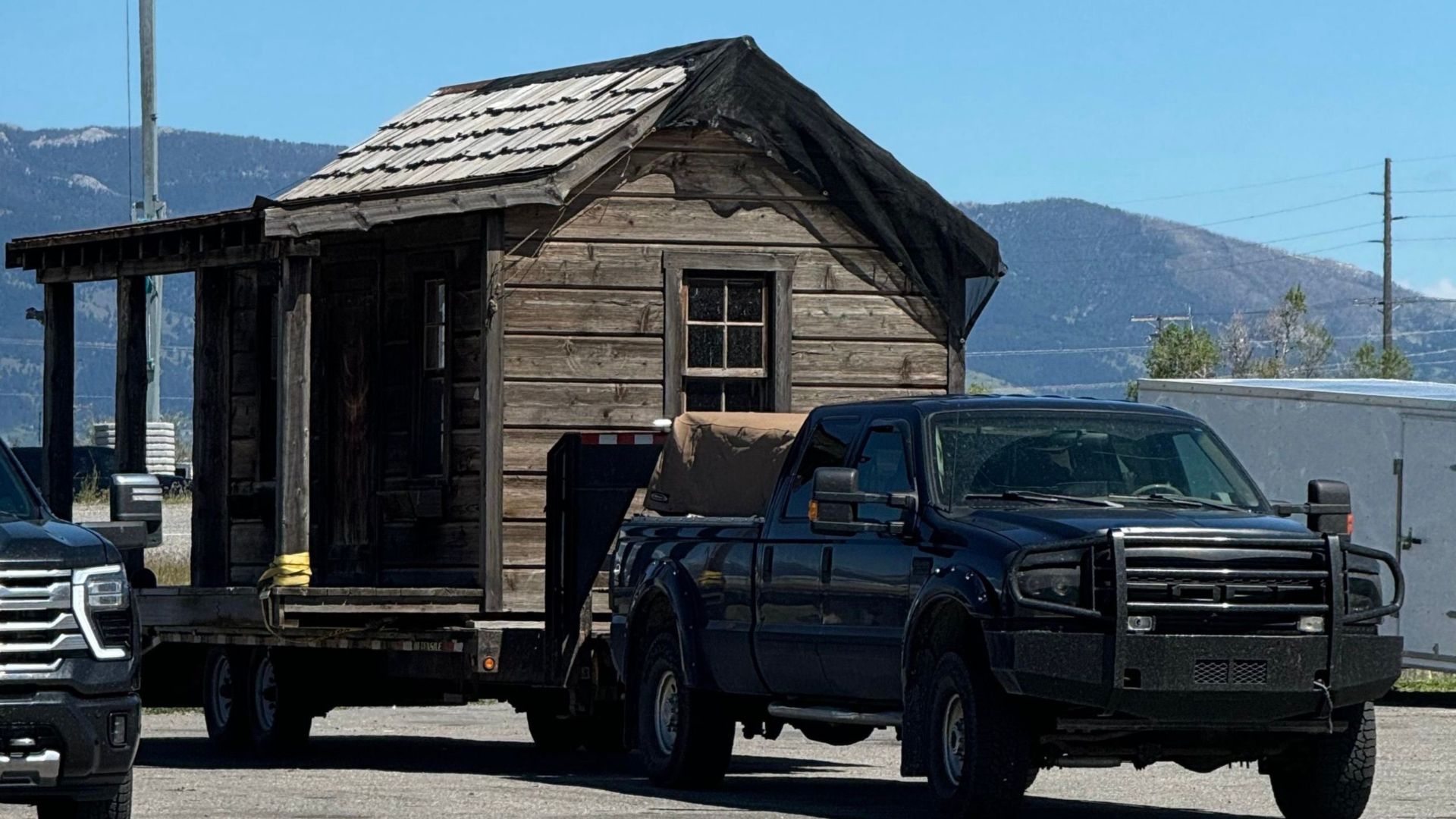 A rustic wooden cabin being towed by a pickup truck on a clear, sunny day.