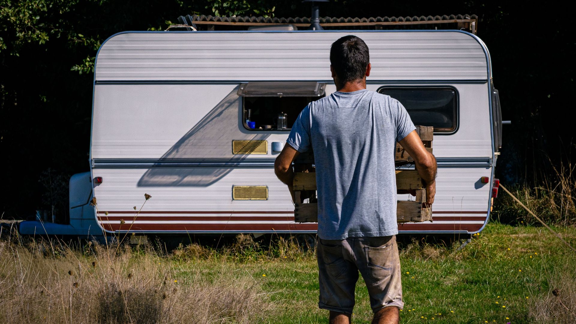 A man carrying a wooden crate approaches a caravan in a sunlit grassy area, evoking a rustic outdoor lifestyle.