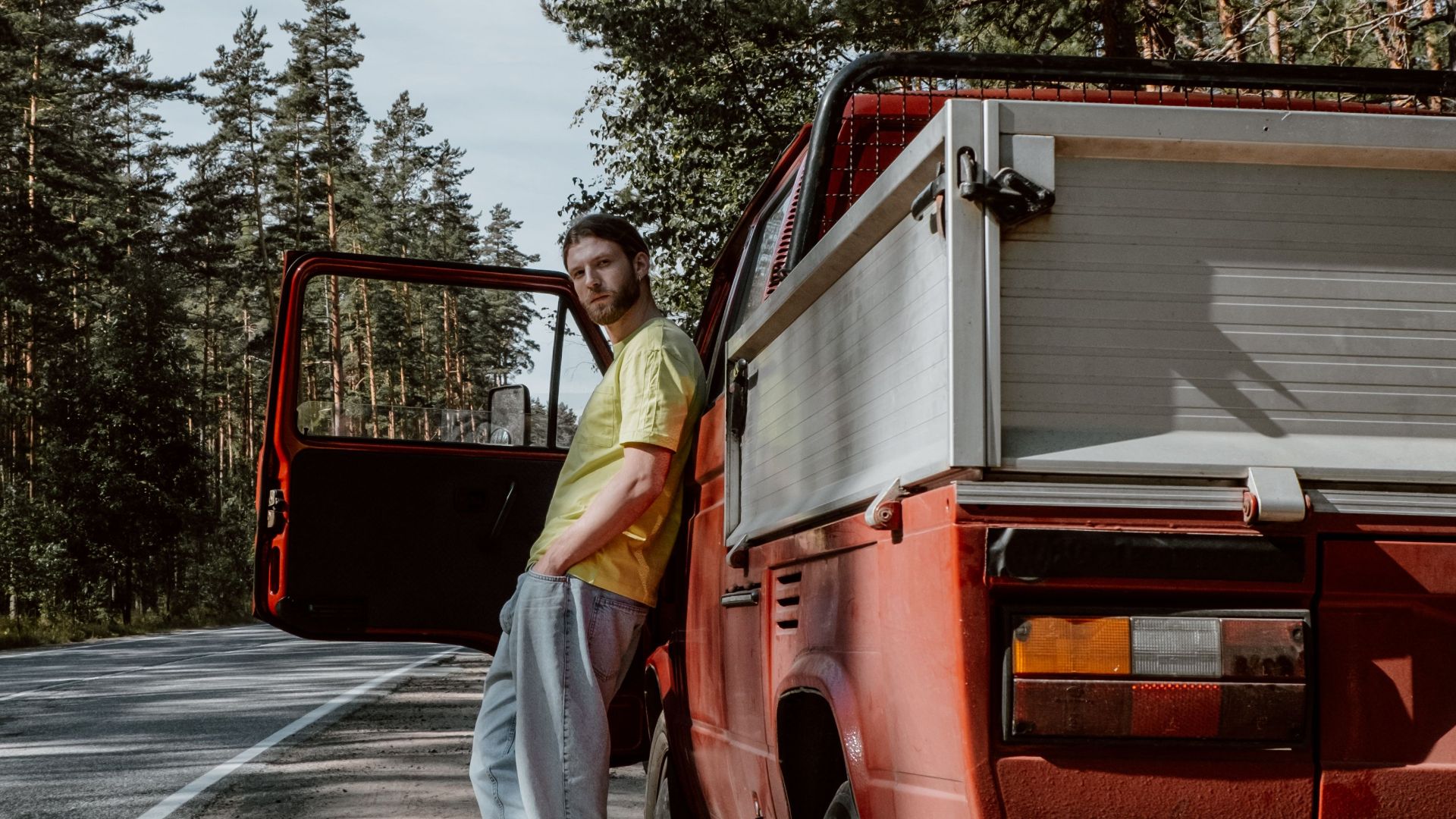 A man stands by his red pickup truck on a quiet country road surrounded by tall trees.