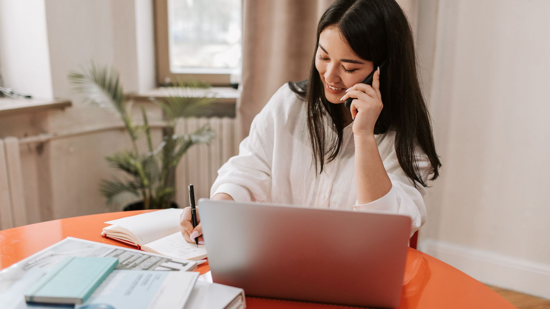 Young woman multitasking with laptop and phone in a modern home office setting.