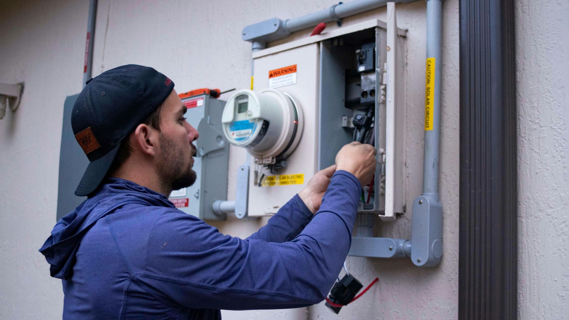 A person in a blue hoodie and baseball cap working on an electrical meter