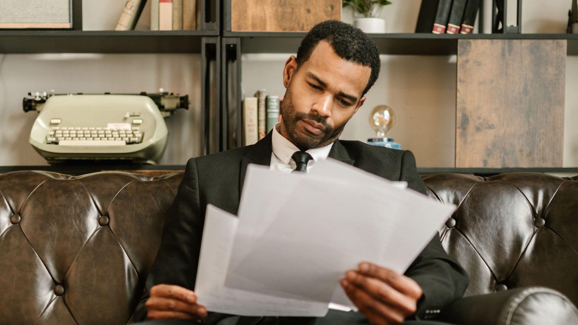 Adult man in a suit reading paperwork on a leather couch in a stylish office setting.