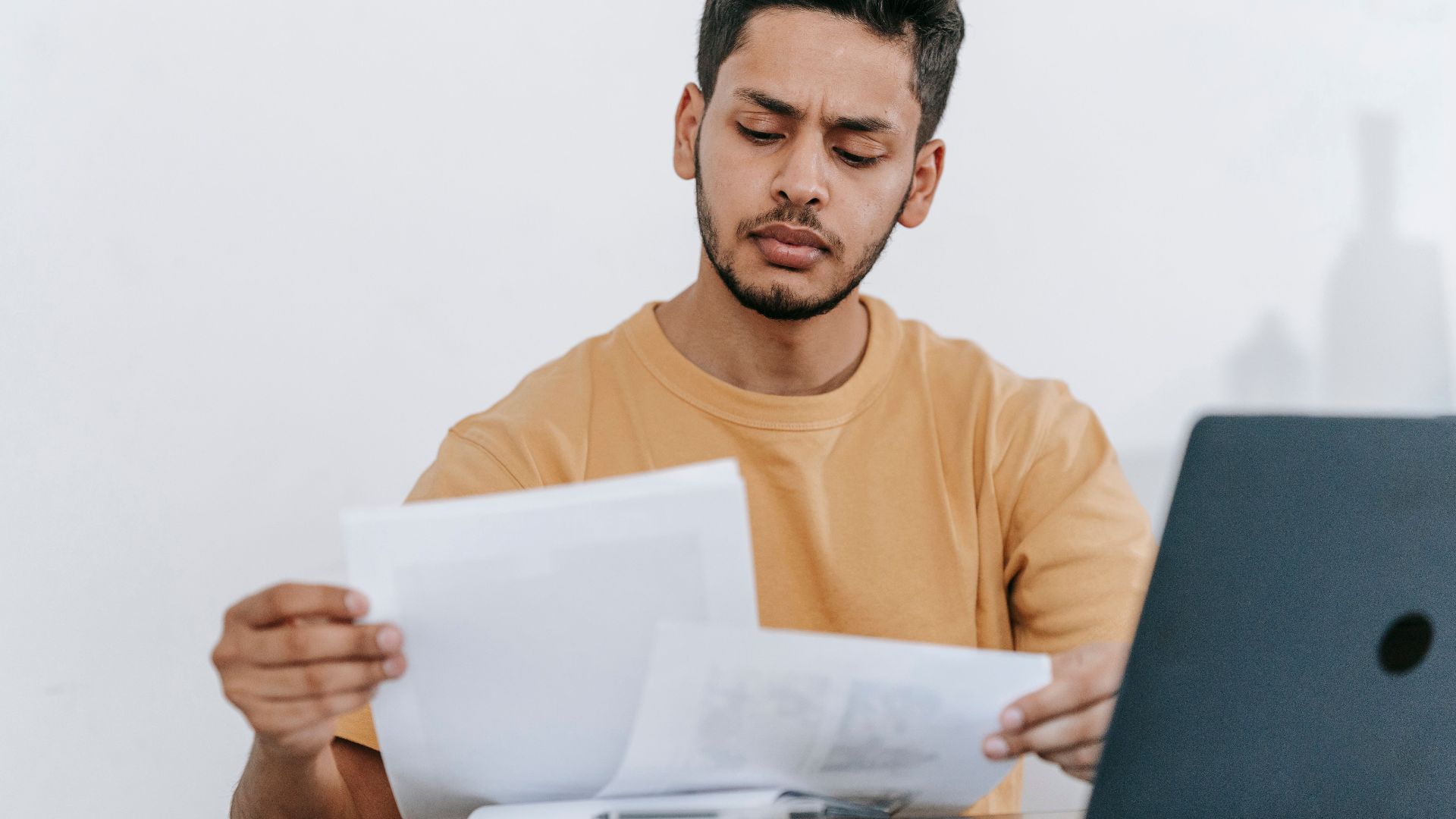 Attentive young bearded Hispanic male remote worker examining papers with results of project while working with laptop in home office