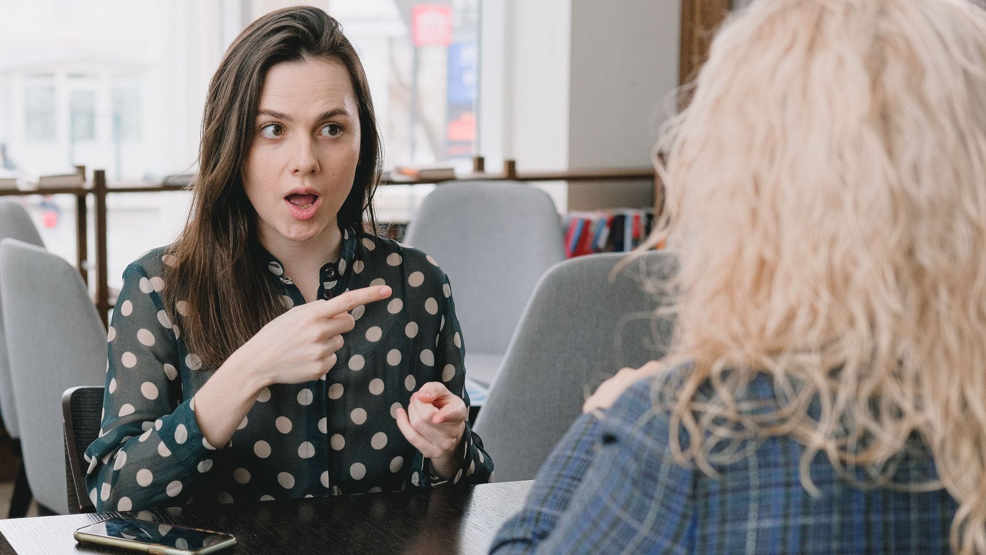 Astonished female pointing away while sitting at table with smartphone and tablet and talking to woman
