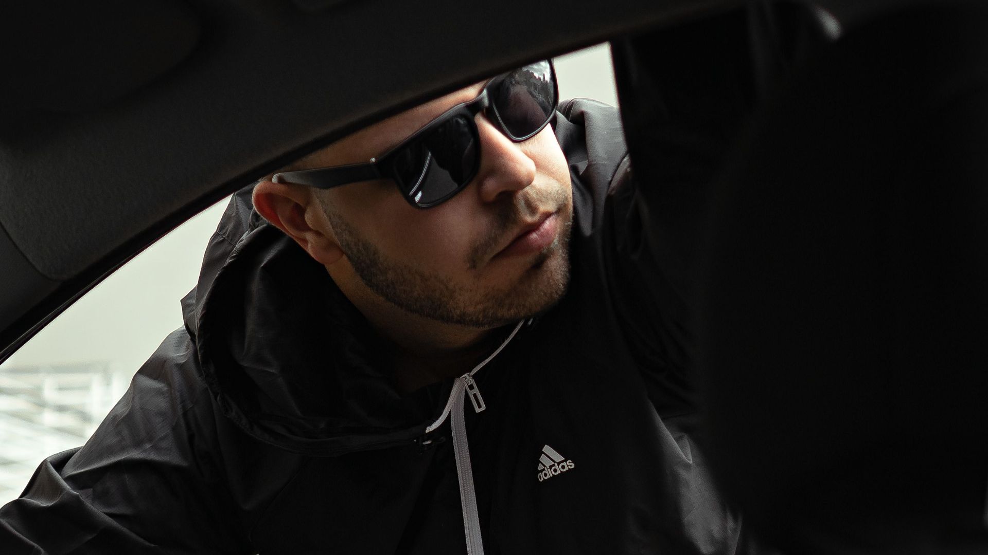 Man wearing sunglasses and jacket looking into a car through the window, creating a mysterious atmosphere.