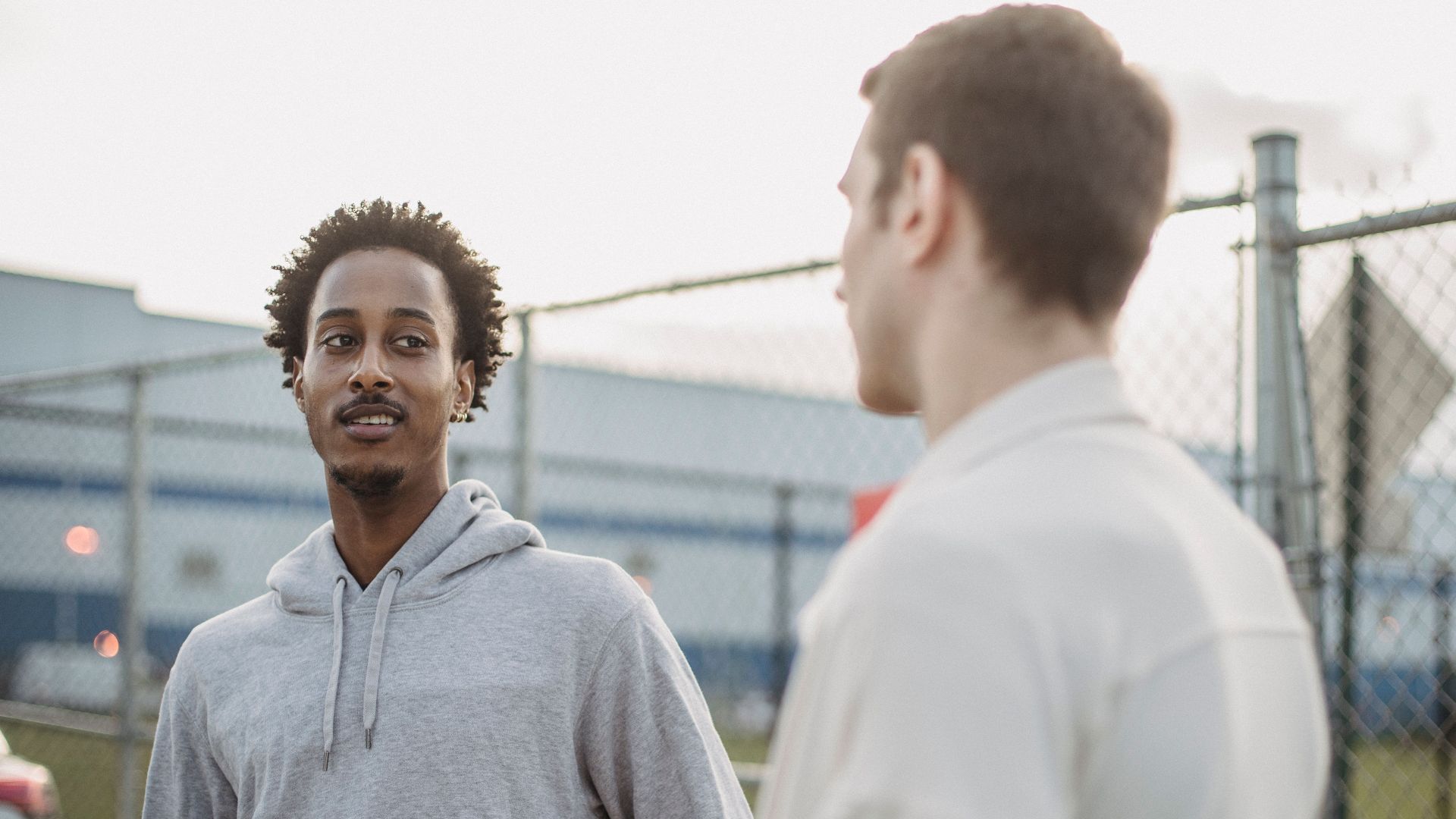 African American male standing with friend against metal grid near car on parking lot