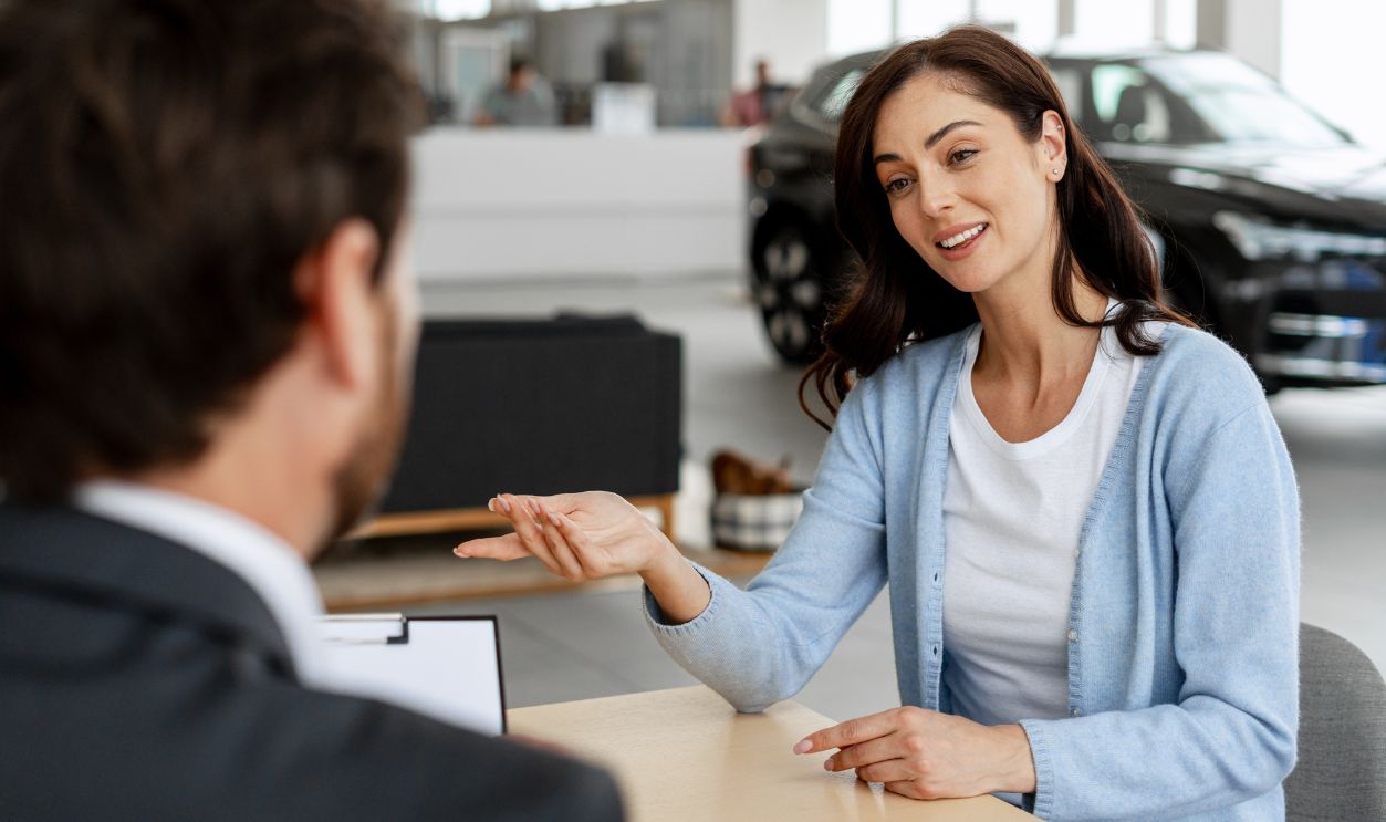 Customer discussing her needs with a car dealer while seated at a desk in a bustling dealership, navigating the buying process together