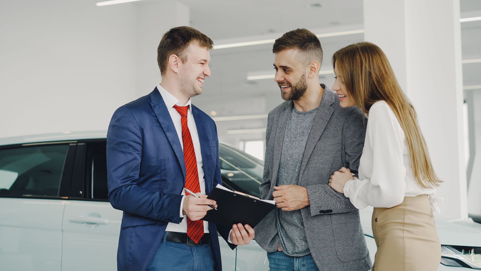 A smiling couple buys a new car from a confident salesman inside a modern car dealership.