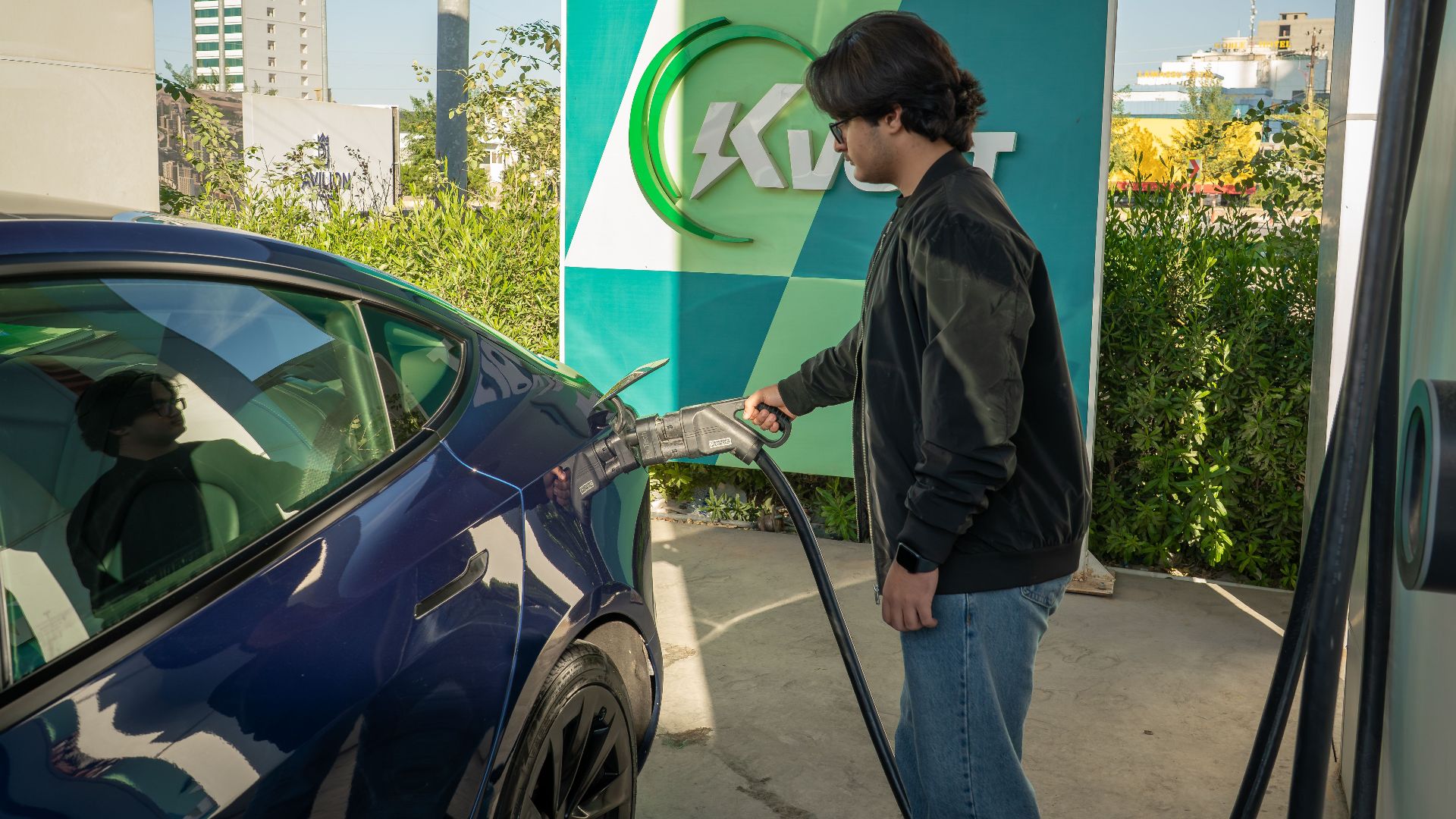 Young man charging a Tesla Model 3 at an outdoor electric vehicle station during the day.